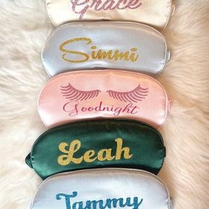 Op de afbeelding: Vijf satijnen slaapmaskers in verschillende kleuren: champagne, zilver, roze, smaragdgroen en wit. Elk masker heeft een naam of zin in glitterletters: Grace, Simmi, Goodnight, Leah en Tammy. De maskers liggen op een zachte witte ondergrond.