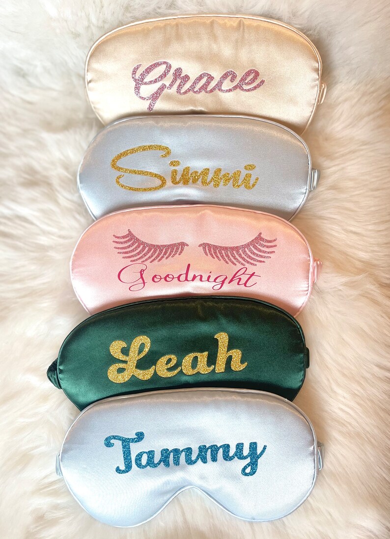 Op de afbeelding: Vijf satijnen slaapmaskers in verschillende kleuren: champagne, zilver, roze, smaragdgroen en wit. Elk masker heeft een naam of zin in glitterletters: Grace, Simmi, Goodnight, Leah en Tammy. De maskers liggen op een zachte witte ondergrond.