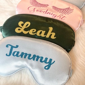 Op de afbeelding: Vier satijnen slaapmaskers in verschillende kleuren: zilver, roze en donkergroen. Elk masker heeft een naam of zin in glitterletters. Het zilveren masker zegt "Tammy", het roze masker zegt "Goodnight", het donkergroene masker zegt "Leah" en het zilveren masker zegt "Simmi".