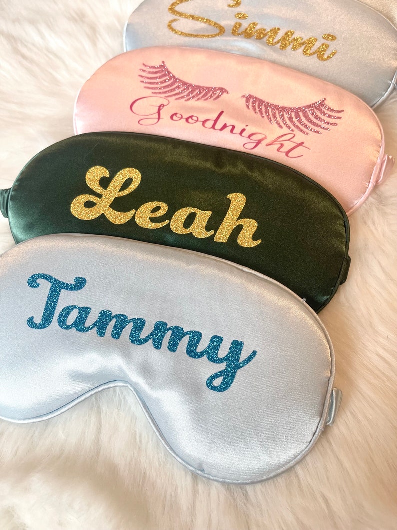 Op de afbeelding: Vier satijnen slaapmaskers in verschillende kleuren: zilver, roze en donkergroen. Elk masker heeft een naam of zin in glitterletters. Het zilveren masker zegt "Tammy", het roze masker zegt "Goodnight", het donkergroene masker zegt "Leah" en het zilveren masker zegt "Simmi".