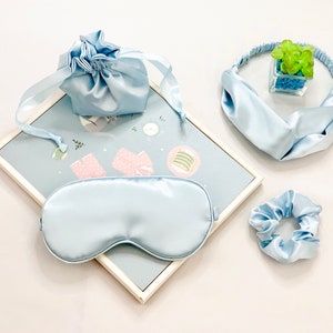 Op de afbeelding: Een lichtblauwe satijnen slaapset met een oogmasker, scrunchie, hoofdband en trekkoordtas. De set is gerangschikt op een lichtblauw oppervlak met een patroon, met een kleine decoratieve plant in een glazen kubus.