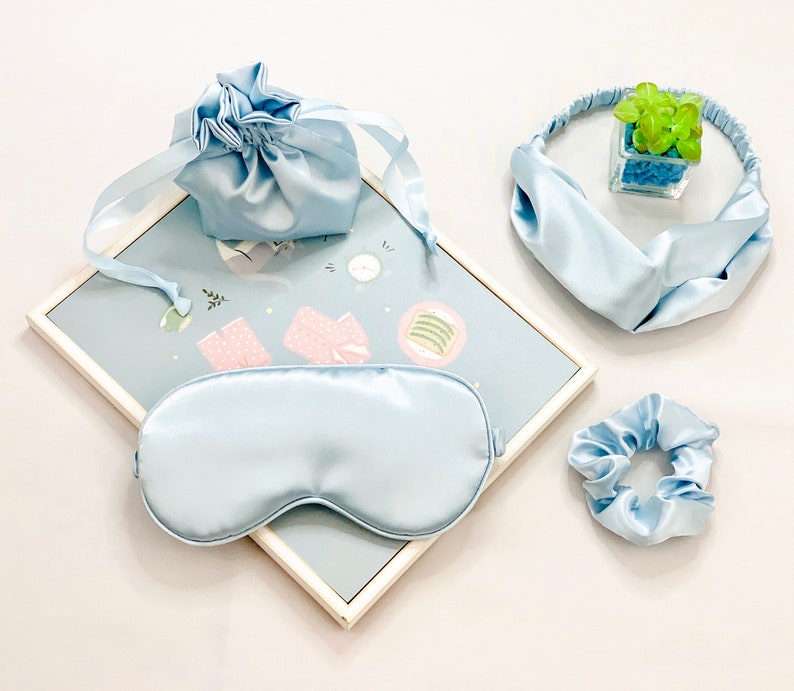 Op de afbeelding: Een lichtblauwe satijnen slaapset met een oogmasker, scrunchie, hoofdband en trekkoordtas. De set is gerangschikt op een lichtblauw oppervlak met een patroon, met een kleine decoratieve plant in een glazen kubus.