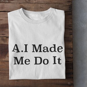 Puede incluir: Camiseta blanca con texto negro que dice "A.I Made Me Do It". La camiseta está doblada y colocada sobre una superficie de madera.