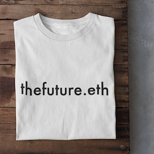 May include: White t-shirt with the text "thefuture.eth" printed in black on the front. The shirt is folded and laid on a wooden surface.