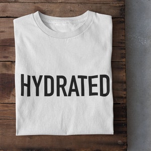 May include: White t-shirt with the word "HYDRATED" printed in black on the front. The t-shirt is folded and laid on a wooden surface. Two white sneakers are on a gray surface below the wooden surface.