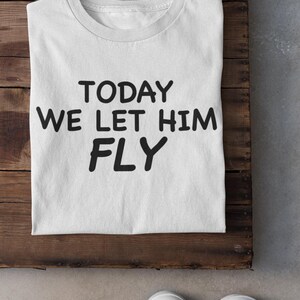 Pode incluir: Camiseta branca com o texto "TODAY WE LET HIM FLY" em letras pretas em negrito. A camiseta está colocada sobre uma superfície de madeira, com um par de tênis brancos na parte inferior.