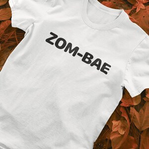 Puede incluir: Camiseta blanca con el texto "ZOM-BAE" impreso en negro, en ángulo en la parte delantera. La camiseta se muestra sobre una superficie con hojas de otoño.
