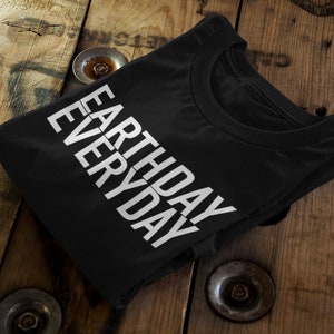 Puede incluir: Camiseta negra con texto blanco que dice "Earth Day Everyday". La camiseta está doblada y sobre una superficie de madera.