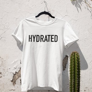 May include: White t-shirt with black text that reads "HYDRATED" hanging on a hanger against a white wall with a cactus in the background.