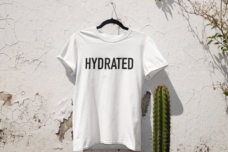 May include: White t-shirt with black text that reads "HYDRATED" hanging on a hanger against a white wall with a cactus in the background.