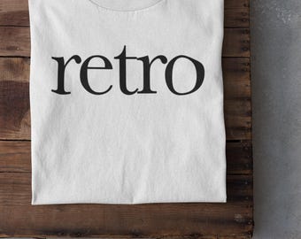 May include: White t-shirt with the word "retro" printed in black. The shirt is laid on a wooden surface, with a pair of white sneakers visible at the bottom. The shirt has a crew neck.