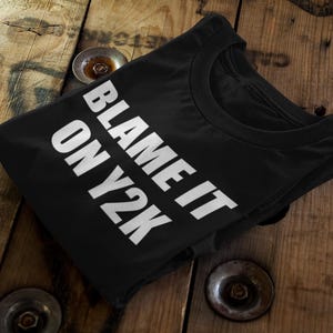 Op de afbeelding: Zwart T-shirt met de tekst "BLAME IT ON Y2K" in witte blokletters. Het shirt is opgevouwen en ligt op een houten oppervlak met metalen accenten.