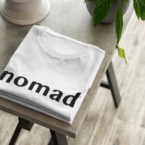 May include: A white t-shirt folded on a gray table with the word "nomad" printed in black on the front.