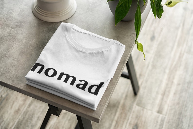 May include: A white t-shirt folded on a gray table with the word "nomad" printed in black on the front.
