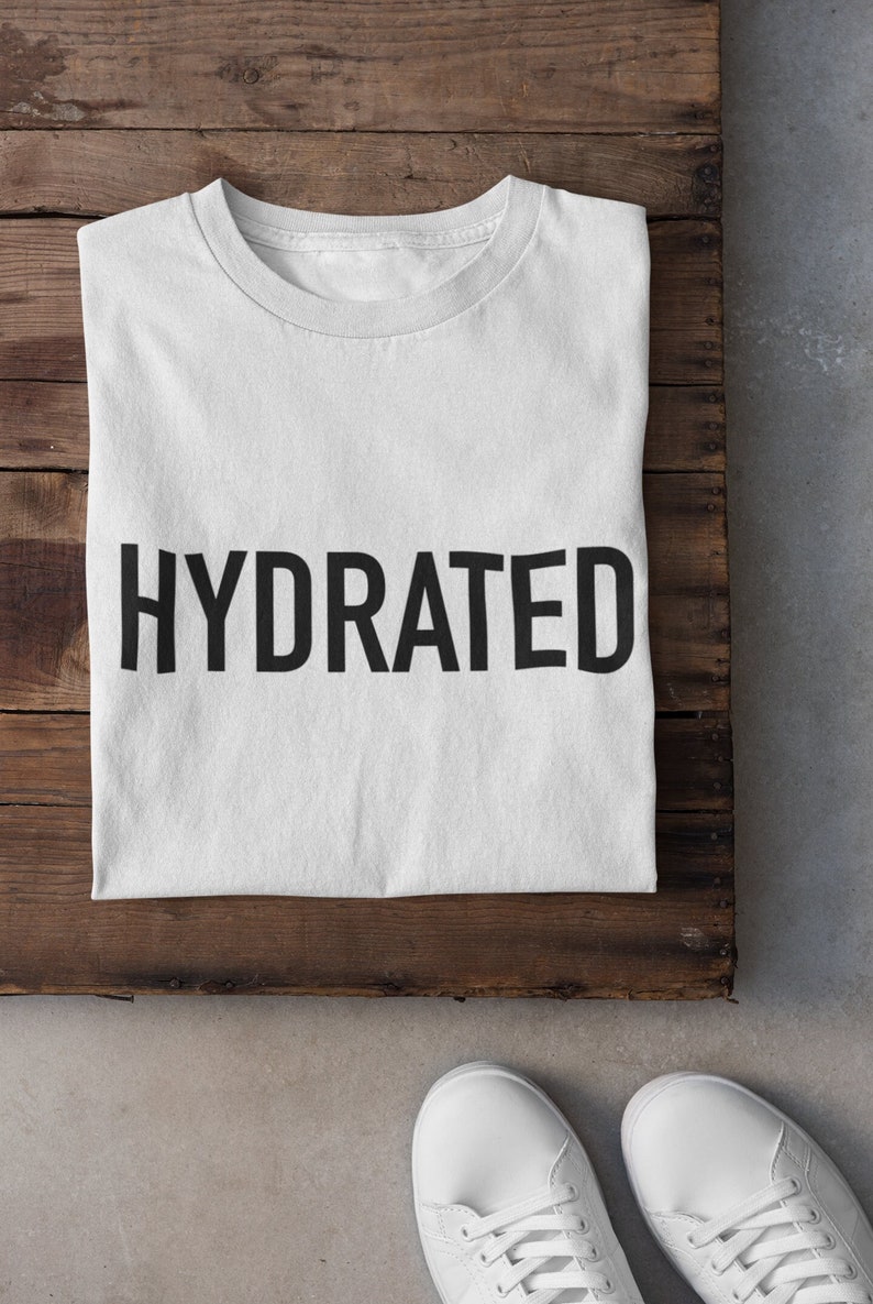 May include: White t-shirt with the word "HYDRATED" printed in black on the front. The t-shirt is folded and laid on a wooden surface. Two white sneakers are on a gray surface below the wooden surface.