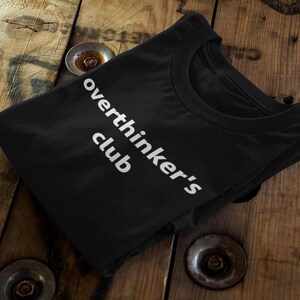 May include: Black t-shirt with the white text "overthinker's club" printed on the front. The shirt is folded and resting on a wooden surface with metal accents.