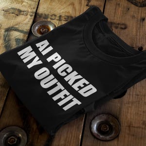 Könnte beinhalten: Schwarzes T-Shirt mit dem weißen Text "AI PICKED MY OUTFIT", auf einer Holzoberfläche. Das T-Shirt ist gefaltet und zeigt den Rundhalsausschnitt. Die Holzoberfläche hat ein rustikales Aussehen.