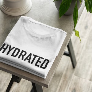 May include: A white t-shirt folded on a gray table with the word "HYDRATED" printed in black on the front.