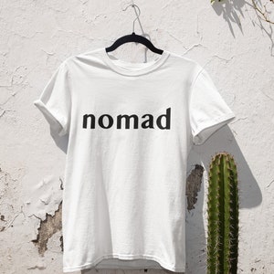 May include: White t-shirt with black text that reads "nomad" hanging on a hanger against a white wall with a cactus in the background.