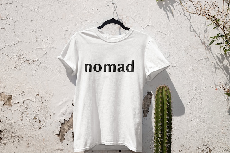 May include: White t-shirt with black text that reads "nomad" hanging on a hanger against a white wall with a cactus in the background.