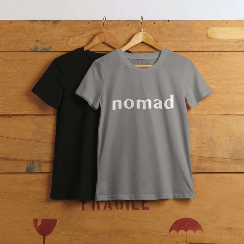 May include: Two t-shirts hanging on wooden hangers. The shirt on the left is black. The shirt on the right is gray with the word "nomad" printed in white on the front.