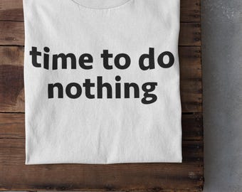 May include: A white t-shirt with the text "time to do nothing" in black lettering. The shirt is laid on a wooden surface, with white trainers visible at the bottom, creating a relaxed vibe.