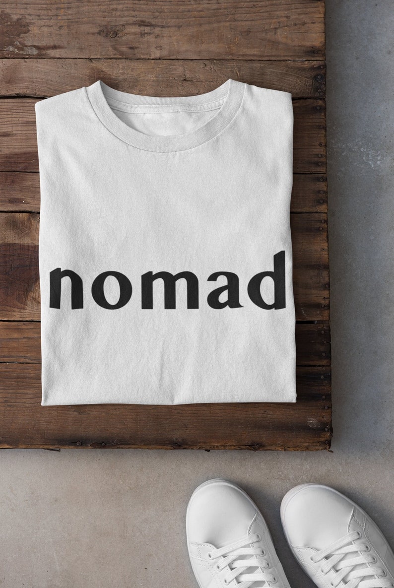 May include: White t-shirt with the word "nomad" printed in black on a wooden background.