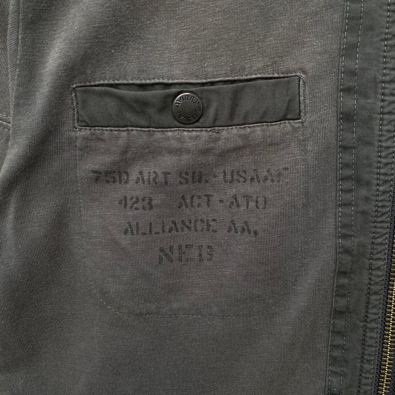 May include: A close-up of a dark gray jacket with a pocket on the left side. The pocket has a black button and the text "750 ART SIL USA", "123 ACT-ATO", "ALLIANCE AA", and "N&deg; 13" printed on it.
