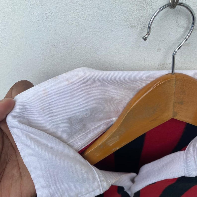 May include: Close-up of a white collared shirt with a red and black striped pattern. The shirt is hanging on a wooden hanger with a metal hook. The collar has some discoloration.