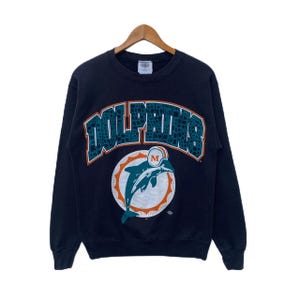 Puede incluir: Sudadera azul marino con la palabra "Dolphins" en letras de bloque turquesa y naranja. Debajo, un logotipo circular con un delfín blanco y turquesa saltando, delineado en naranja y blanco.
