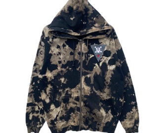 May include: Black and tan tie-dye zip-up hoodie with a hood and front pockets. Features a small patch on the chest with a skull and crossbones design, a red star, and the letter "S".