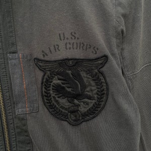 May include: A gray cotton jacket with a black embroidered patch that says "U.S. AIR CORPS" and features a stylized eagle with wings spread.