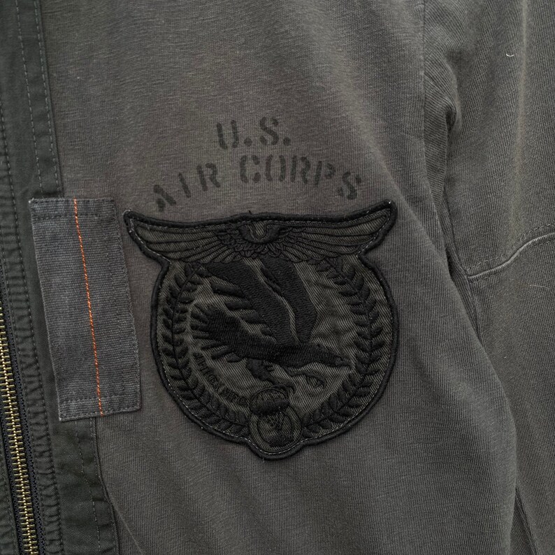 May include: A gray cotton jacket with a black embroidered patch that says "U.S. AIR CORPS" and features a stylized eagle with wings spread.