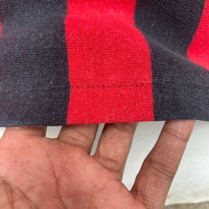 May include: Close-up of a red and navy blue striped fabric. The fabric is a knit material with visible texture. The stripes are wide and evenly spaced. The bottom edge of the fabric shows a seam with visible stitching.