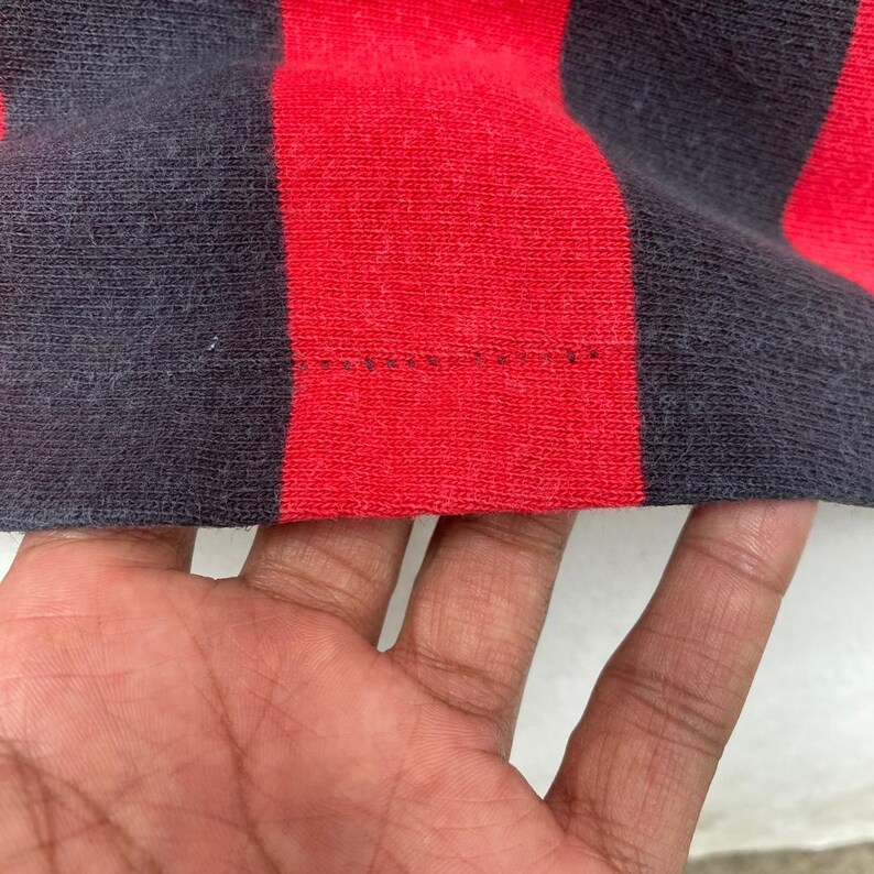 May include: Close-up of a red and navy blue striped fabric. The fabric is a knit material with visible texture. The stripes are wide and evenly spaced. The bottom edge of the fabric shows a seam with visible stitching.