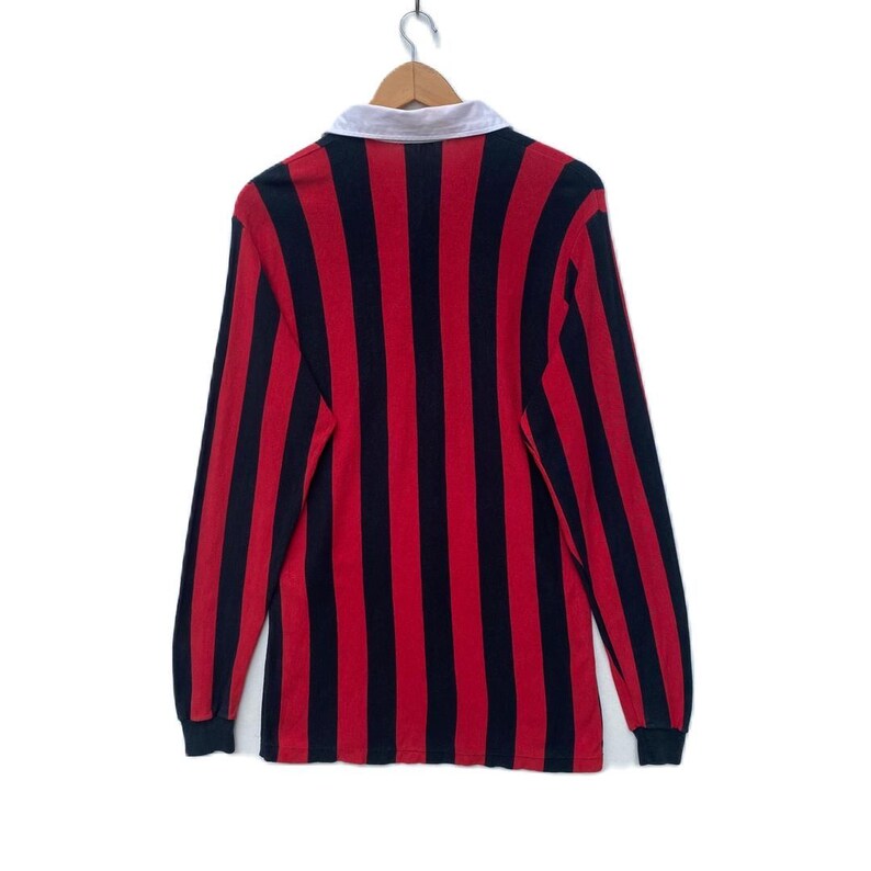 May include: A long-sleeved rugby shirt with vertical red and navy blue stripes. The shirt has a white collar and is hanging on a wooden hanger. The cuffs are navy blue.