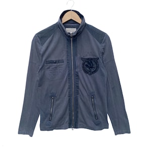 May include: A blue zip-up jacket with a U.S. Air Corps logo on the front. The jacket has a stand-up collar and two zippered pockets on the sides.