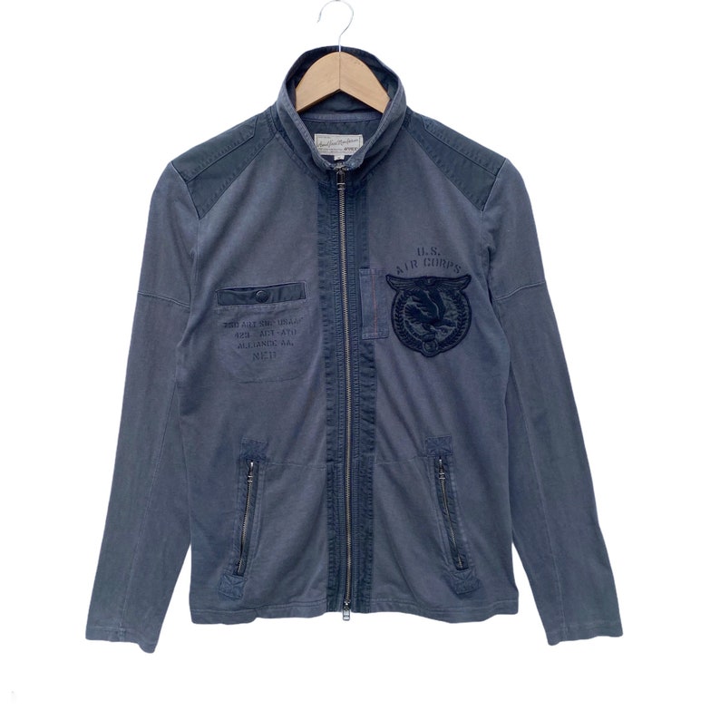 May include: A blue zip-up jacket with a U.S. Air Corps logo on the front. The jacket has a stand-up collar and two zippered pockets on the sides.