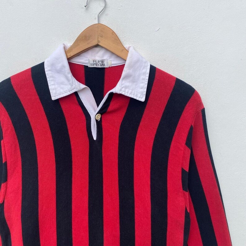 May include: A red and black striped long-sleeve shirt with a white collar. The shirt has a button-down placket and the text "FLIPS SPECIAL" is visible on the collar. The shirt is hanging on a wooden hanger.