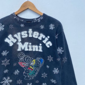 K&ouml;nnte beinhalten: Dunkelblaues Sweatshirt mit dem wei&szlig;en Schriftzug "Hysteric Mini". Das Sweatshirt hat eine Cartoon-Grafik und Schneeflocken-Designs. Der Kragen hat ein wei&szlig;es Schneeflockenmuster. Das Sweatshirt h&auml;ngt an einem Holzb&uuml;gel.