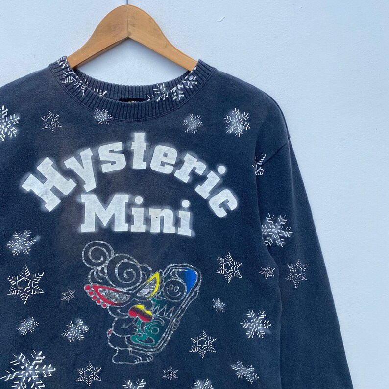 K&ouml;nnte beinhalten: Dunkelblaues Sweatshirt mit dem wei&szlig;en Schriftzug "Hysteric Mini". Das Sweatshirt hat eine Cartoon-Grafik und Schneeflocken-Designs. Der Kragen hat ein wei&szlig;es Schneeflockenmuster. Das Sweatshirt h&auml;ngt an einem Holzb&uuml;gel.