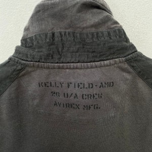 May include: A dark gray cotton jacket with a corduroy collar. The back of the jacket has the text "KELLY FIELD - AND 28 U.S.A. CREW AVIREX INC." printed in black.
