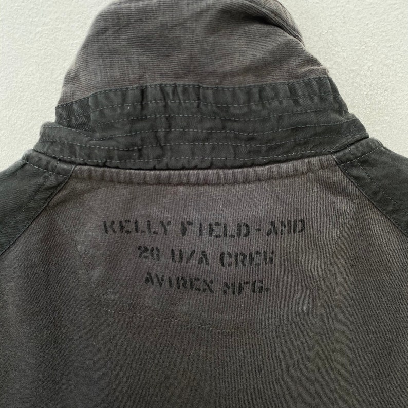 May include: A dark gray cotton jacket with a corduroy collar. The back of the jacket has the text "KELLY FIELD - AND 28 U.S.A. CREW AVIREX INC." printed in black.