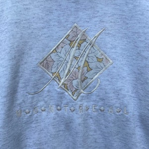 May include: A light gray sweatshirt with a white embroidered logo of a stylized "M" with a floral pattern inside a diamond shape. The text "MONTREAL" is embroidered below the logo with a maple leaf between each letter.