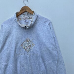 May include: A gray sweatshirt with a floral print on the inside of the collar. The front of the sweatshirt features a diamond-shaped logo with a floral design and the text "MOUNTAIN EAGLE".