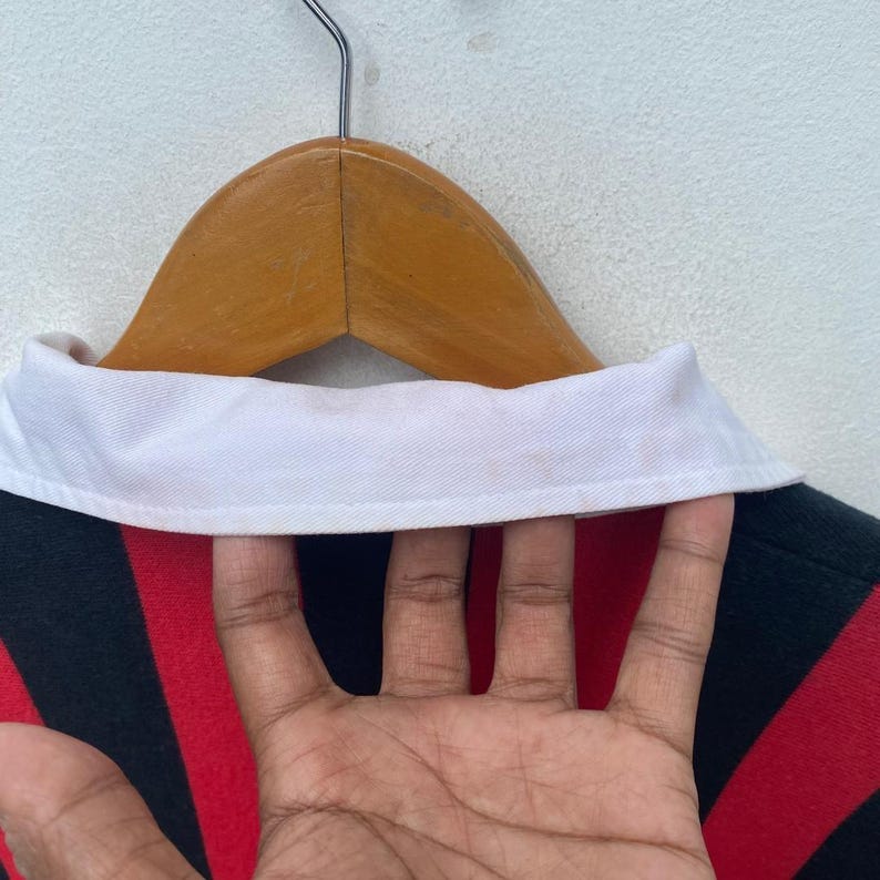 May include: Close-up of a striped rugby shirt hanging on a wooden hanger. The shirt features alternating red and black stripes with a white collar. The shirt is made of a thick, durable material.