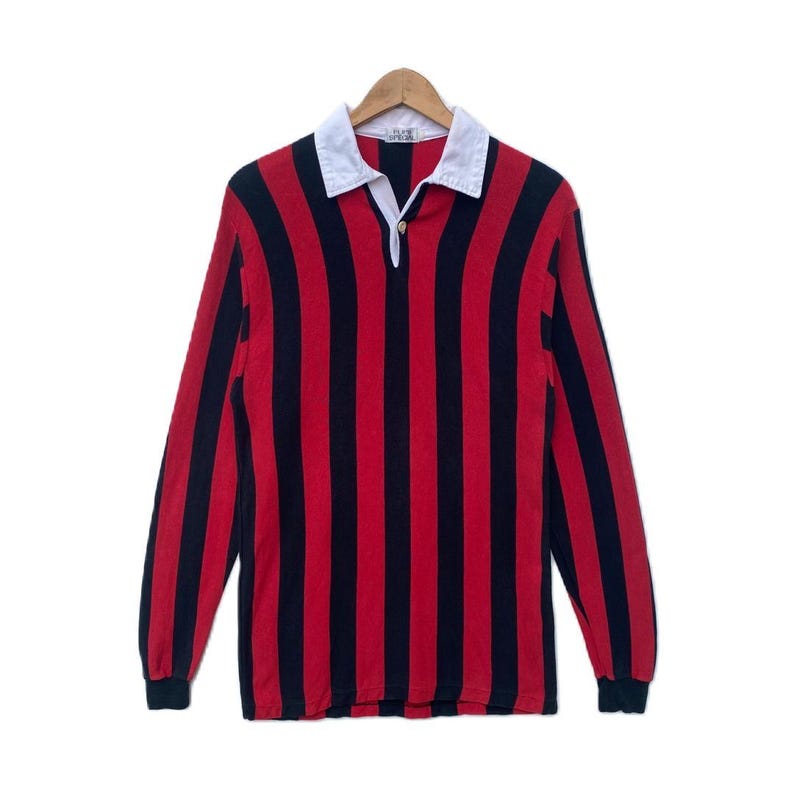 May include: A long-sleeved rugby shirt with vertical red and black stripes. The shirt has a white collar and a button-up placket. The cuffs are black. The shirt is hanging on a wooden hanger against a white background.