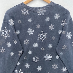 K&ouml;nnte beinhalten: Ein dunkelgraues Sweatshirt mit einem Muster aus wei&szlig;en Schneeflocken. Die Schneeflocken sind unterschiedlich gro&szlig; und &uuml;ber den Stoff verteilt. Das Rundhals-Sweatshirt wird auf einem Holzb&uuml;gel pr&auml;sentiert.