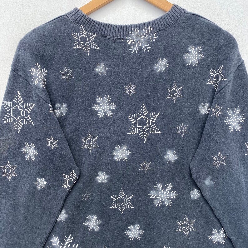 K&ouml;nnte beinhalten: Ein dunkelgraues Sweatshirt mit einem Muster aus wei&szlig;en Schneeflocken. Die Schneeflocken sind unterschiedlich gro&szlig; und &uuml;ber den Stoff verteilt. Das Rundhals-Sweatshirt wird auf einem Holzb&uuml;gel pr&auml;sentiert.