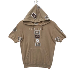 Peut inclure: Un sweat à capuche à manches courtes de couleur beige avec une poche kangourou. Le sweat a le texte "PINK HOUSE" imprimé sur le devant, ainsi que plusieurs motifs de fleurs brodés.